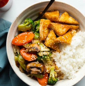 A bowl of steamed rice, tender-crisp vegetables and golden tofu topped with teriyaki sauce and toasted sesame seeds.