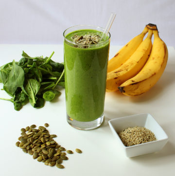 A large glass of Green Protein Power Breakfast Smoothie surrounded by key ingredients: spinach, pumpkin seeds, bananas, hemp seeds.