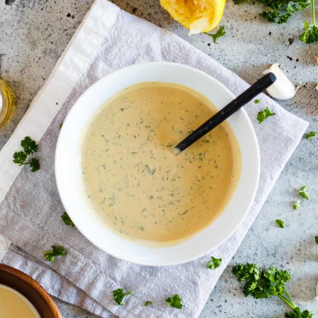 A bowl of Vegan Tahini Goddess Dressing surrounded by key ingredients on a concrete background.