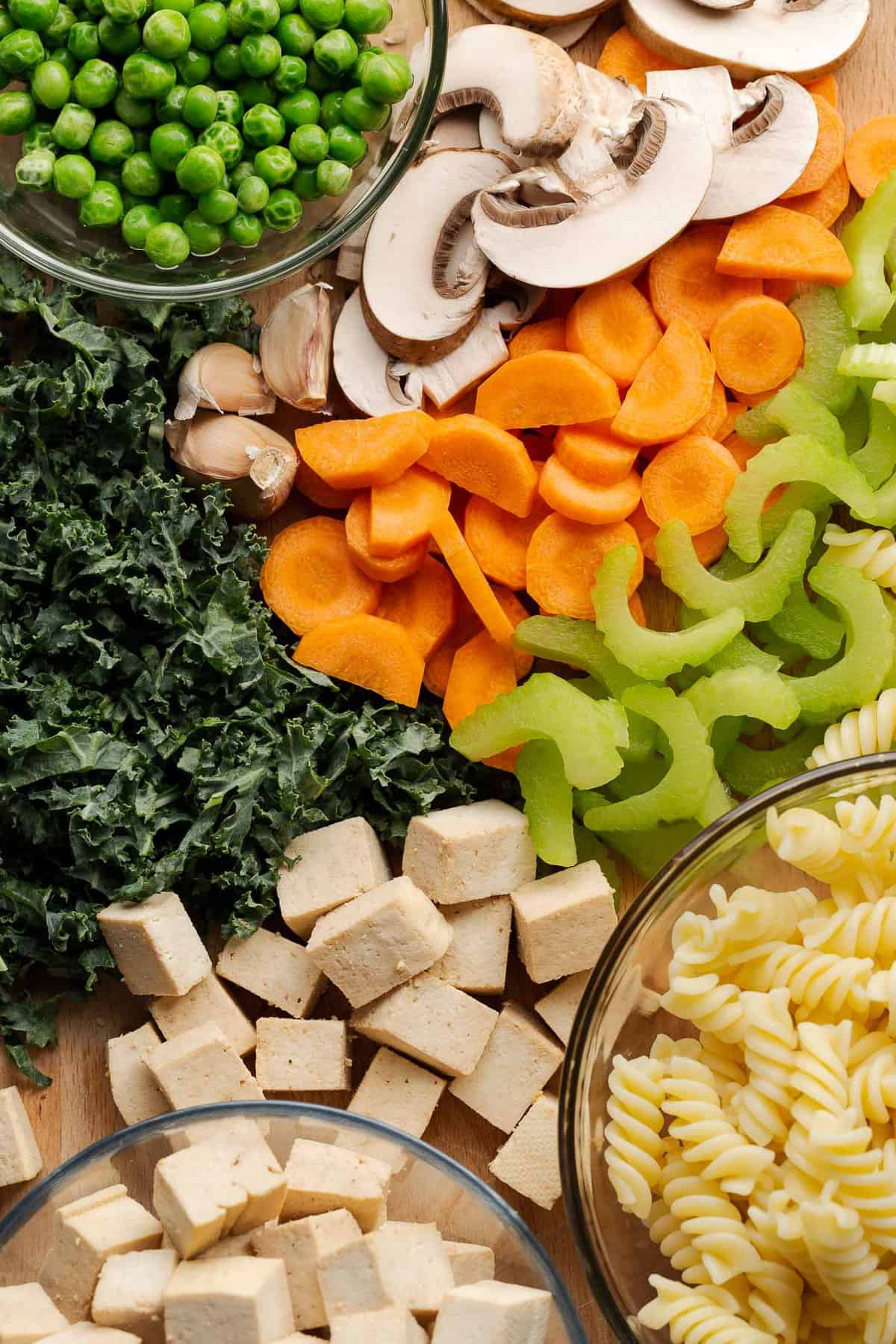 Tofu noodle soup ingredients on a cutting board: peas, mushrooms, kale, garlic, carrot, celery, tofu, and cooked fusili noodles.