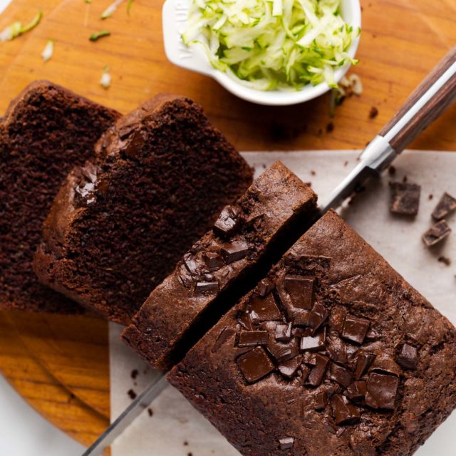 Bird's eye view of a loaf of vegan chocolate zucchini bread being sliced with a wood-handled serrated knife. The loaf is on a small piece of parchment paper on a round wooden teak cutting board on a marble countertop.