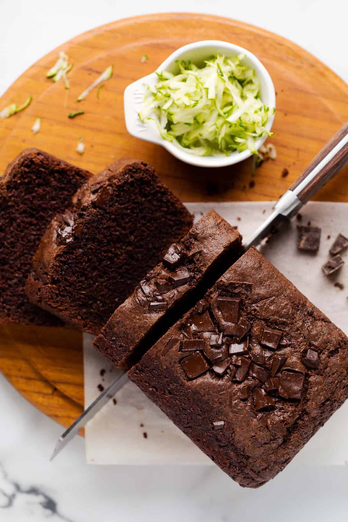 Bird's eye view of a loaf of vegan chocolate zucchini bread being sliced with a wood-handled serrated knife. The loaf is on a small piece of parchment paper on a round wooden teak cutting board on a marble countertop.